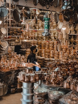A busy market stall filled with a variety of shiny brass and copper kitchenware and decorative items. The items are stacked high on shelves and tables, reflecting the light and creating a warm, metallic ambiance. A person is sitting among the items, partially obscured by the abundance of metallic goods.