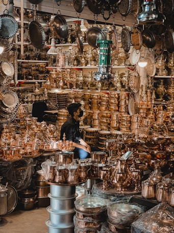 A busy market stall filled with a variety of shiny brass and copper kitchenware and decorative items. The items are stacked high on shelves and tables, reflecting the light and creating a warm, metallic ambiance. A person is sitting among the items, partially obscured by the abundance of metallic goods.