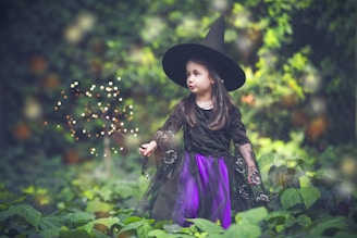 A young girl in a witch costume stands in a lush green garden. She is wearing a black pointed hat and a black dress with purple accents. In her hand, she holds a wand that emits magical sparkles. The background is filled with blurred greenery, creating an enchanting atmosphere.