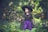 A young girl in a witch costume stands in a lush green garden. She is wearing a black pointed hat and a black dress with purple accents. In her hand, she holds a wand that emits magical sparkles. The background is filled with blurred greenery, creating an enchanting atmosphere.