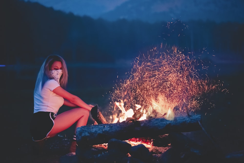 a woman sitting in front of a campfire wearing a face mask