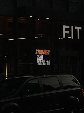 A dark street scene featuring an illuminated gym sign with the motivational phrase 'STRONGER THAN YESTERDAY' in bright neon colors. In front of the building, a black vehicle is parked. The sign on the building reads 'FIT' in bold, white letters.