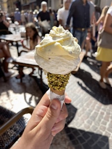 A hand is holding an ice cream cone topped with two scoops of light yellow gelato, possibly lemon or vanilla flavored. The cone is coated with crushed pistachios, adding a vibrant green texture. In the background, there is a busy outdoor setting with people walking and others sitting at tables, suggesting a lively street or plaza atmosphere.