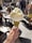 A hand is holding an ice cream cone topped with two scoops of light yellow gelato, possibly lemon or vanilla flavored. The cone is coated with crushed pistachios, adding a vibrant green texture. In the background, there is a busy outdoor setting with people walking and others sitting at tables, suggesting a lively street or plaza atmosphere.