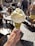 A hand is holding an ice cream cone topped with two scoops of light yellow gelato, possibly lemon or vanilla flavored. The cone is coated with crushed pistachios, adding a vibrant green texture. In the background, there is a busy outdoor setting with people walking and others sitting at tables, suggesting a lively street or plaza atmosphere.