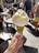 A hand is holding an ice cream cone topped with two scoops of light yellow gelato, possibly lemon or vanilla flavored. The cone is coated with crushed pistachios, adding a vibrant green texture. In the background, there is a busy outdoor setting with people walking and others sitting at tables, suggesting a lively street or plaza atmosphere.