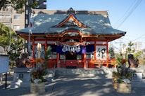 A traditional Shinto shrine with a sloping roof and vibrant red accents stands at the center. The entrance is flanked by two stone guardian statues and adorned with decorative elements. The foreground features ornate arrangements of bamboo and flowers, suggesting a festive or ceremonial occasion. The Japanese flag is visible to the left, and modern buildings are partially seen in the background, juxtaposing the traditional and contemporary.
