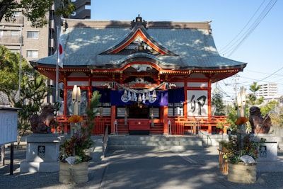 A traditional Shinto shrine with a sloping roof and vibrant red accents stands at the center. The entrance is flanked by two stone guardian statues and adorned with decorative elements. The foreground features ornate arrangements of bamboo and flowers, suggesting a festive or ceremonial occasion. The Japanese flag is visible to the left, and modern buildings are partially seen in the background, juxtaposing the traditional and contemporary.