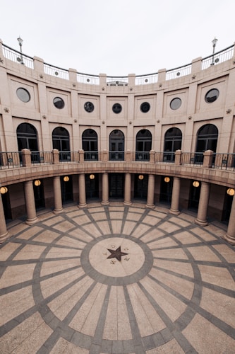 A grand architectural structure forms a circular courtyard surrounded by a series of columns and arched windows. The courtyard features a geometric tiled pattern on the ground with a large central star design. The upper level includes a balcony with a railing and ornate details along the facade.