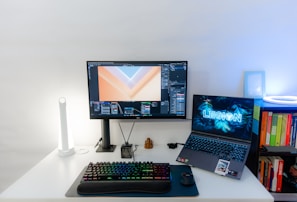 A modern, organized workspace featuring a large monitor displaying graphic design software alongside a laptop with a vibrant logo. A sleek keyboard and mouse are positioned on the desk, which also holds small decorative items and a desk lamp. A bookshelf with colorful books is visible nearby.