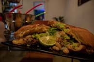 A sliced sandwich on a black tray is filled with various ingredients including mashed avocado, lettuce, beans, chopped onions, and a visible slice of lime. The background features metal buckets with colorful straws and a blurred setting suggesting a restaurant or café environment.