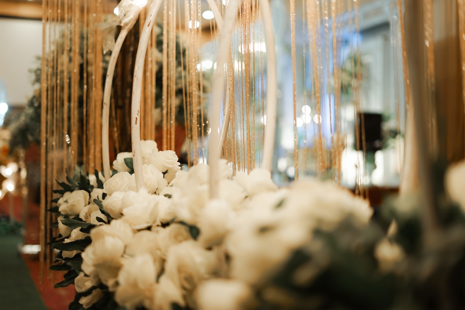 A bouquet of white flowers, possibly roses or peonies, is arranged in an elegant display with vertical gold strands hanging around it. The setting highlights a sophisticated and luxurious atmosphere, likely in an indoor space such as a venue or event hall. Warm lighting enhances the ambiance, with blurred lights visible in the background, contributing to a romantic and festive mood.