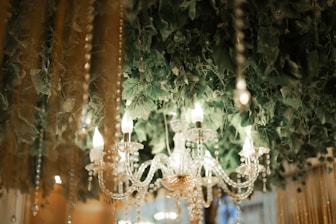 A crystal chandelier with lit candles hanging amidst lush green leaves and decorative strings of beads. The chandelier's intricate design reflects the surrounding warm light, creating an elegant and sophisticated ambiance.