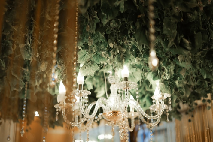 A crystal chandelier with lit candles hanging amidst lush green leaves and decorative strings of beads. The chandelier's intricate design reflects the surrounding warm light, creating an elegant and sophisticated ambiance.