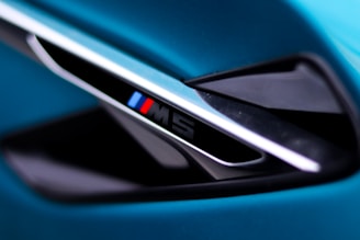 A close-up view of a sleek automotive detail, featuring a diagonal vent with an emblem displaying the letter 'M' alongside blue and red stripes. The surrounding surfaces have a glossy black and metallic finish, set against a vibrant teal background.