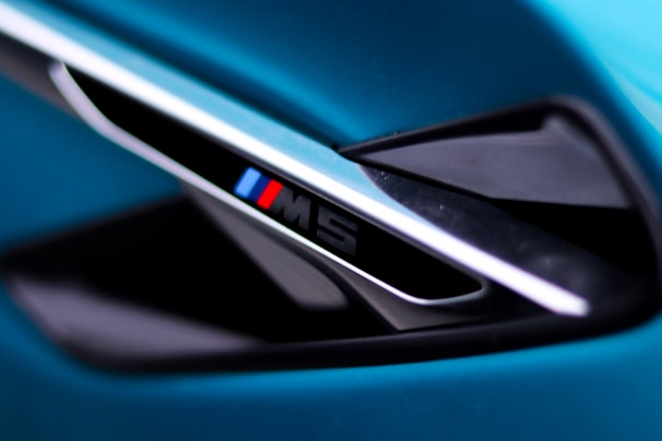 A close-up view of a sleek automotive detail, featuring a diagonal vent with an emblem displaying the letter 'M' alongside blue and red stripes. The surrounding surfaces have a glossy black and metallic finish, set against a vibrant teal background.