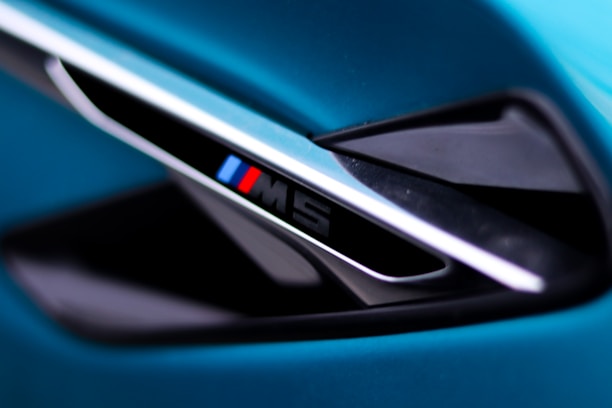 A close-up view of a sleek automotive detail, featuring a diagonal vent with an emblem displaying the letter 'M' alongside blue and red stripes. The surrounding surfaces have a glossy black and metallic finish, set against a vibrant teal background.
