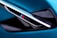 A close-up view of a sleek automotive detail, featuring a diagonal vent with an emblem displaying the letter 'M' alongside blue and red stripes. The surrounding surfaces have a glossy black and metallic finish, set against a vibrant teal background.