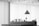 A monochrome image of an interior space featuring a modern hanging pendant light above a shelf or desk. Several small objects are neatly arranged along the surface, including bottles, vases with plants, and a sign. The ceiling has an array of circular cutouts, and the large windows in the background are covered with blinds or curtains, diffusing the light.