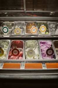 A variety of ice cream flavors displayed in metal containers inside a glass-topped freezer. Each flavor is labeled with circular tags, showing different colors and text. The ice creams include a mix of vibrant colors such as green, purple, and pink.