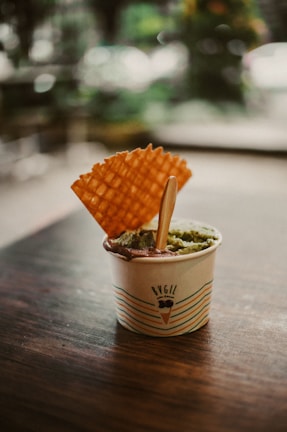A small paper cup filled with gelato sits on a dark wooden table. The cup features colorful stripes and the word 'BuGiL' printed on it. Two flavors of gelato are visible, with a prominent green scoop and a hint of chocolate around the edge. A waffle cookie is inserted upright into the gelato, next to a small wooden spoon.
