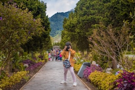 A person is taking a photograph in a lush garden pathway lined with vibrant flowers and large trees. In the distance, a group of people, including children, walk along the pathway. The backdrop features scenic mountains and a partly cloudy sky.
