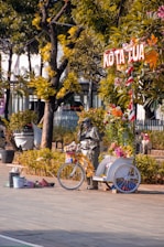 A person painted in silver is standing next to a decorated bicycle with flowers and a bucket for tips in a public square. There are trees and people sitting on benches in the background, and a sign above the bicycle reads 'KOTA TUA'.