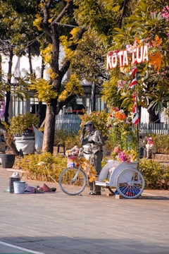 A person painted in silver is standing next to a decorated bicycle with flowers and a bucket for tips in a public square. There are trees and people sitting on benches in the background, and a sign above the bicycle reads 'KOTA TUA'.