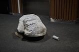 A person wearing a white jacket covered in various handwritten messages is kneeling on the carpeted floor with their head bowed down. Beside them on the floor is an empty aluminum can. The background includes wooden paneling and an elevator door.