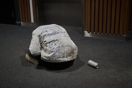 A person wearing a white jacket covered in various handwritten messages is kneeling on the carpeted floor with their head bowed down. Beside them on the floor is an empty aluminum can. The background includes wooden paneling and an elevator door.