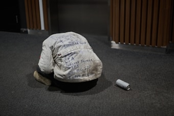 A person wearing a white jacket covered in various handwritten messages is kneeling on the carpeted floor with their head bowed down. Beside them on the floor is an empty aluminum can. The background includes wooden paneling and an elevator door.