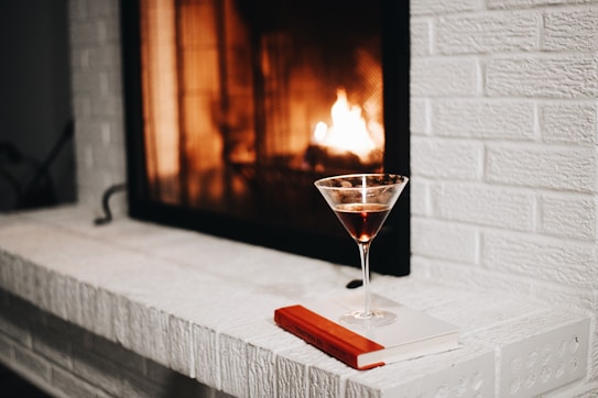 A cozy scene featuring a lit fireplace with a warm, inviting glow. In the foreground, a martini glass filled with a dark liquid rests on a white book with a red spine, placed on the brick hearth.