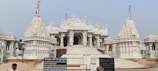 A grand, intricately carved white marble temple with multiple domes and spires, featuring extensive architectural details. The foreground includes some Hindi text on boards, a person, and what appears to be construction material like sand.