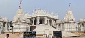 A grand, intricately carved white marble temple with multiple domes and spires, featuring extensive architectural details. The foreground includes some Hindi text on boards, a person, and what appears to be construction material like sand.