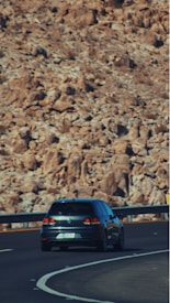 A dark-colored car is driving along a curving highway with a rocky, mountainous landscape in the background. The road appears well-maintained with clear lane markings and a metal guardrail. The scene is likely captured during daytime, suggested by the lighting.