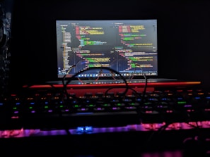 A laptop displaying colorful code on its screen is positioned in a dark environment. The keyboard in the foreground features backlit keys with various neon colors. A pair of glasses rests on the keyboard, casting a shadow over the keys.