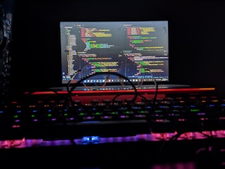 A laptop displaying colorful code on its screen is positioned in a dark environment. The keyboard in the foreground features backlit keys with various neon colors. A pair of glasses rests on the keyboard, casting a shadow over the keys.