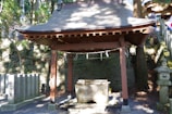 A traditional Japanese purification pavilion stands in a serene forest setting. The structure features a wooden roof supported by four posts, with a stone water basin beneath, used for ritual cleansing. Stone monuments and lanterns surround the area, partially covered by the shadows of nearby trees. Sunlight filters through the canopy, creating a peaceful, dappled light effect.