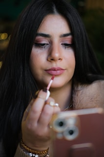 A person with long dark hair is applying lipstick using a lip brush while holding a smartphone in the other hand. The background is blurred, focusing on the facial features and the act of applying makeup.