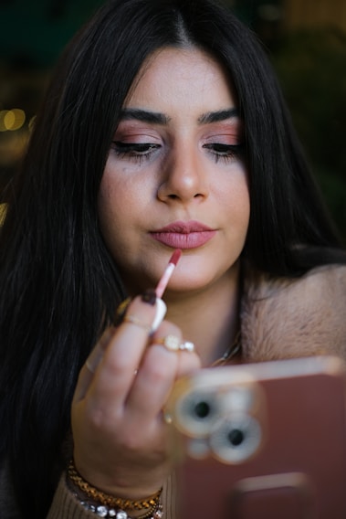 A person with long dark hair is applying lipstick using a lip brush while holding a smartphone in the other hand. The background is blurred, focusing on the facial features and the act of applying makeup.