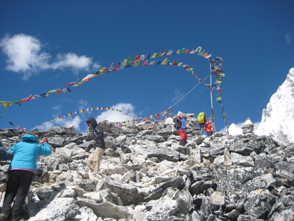 A rocky mountain terrain with several people climbing, surrounded by vibrant prayer flags fluttering in the wind. The sky is clear and blue, and snow can be seen on the right edge of the image.