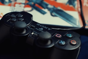 A close-up of a PlayStation controller with visible buttons, including the classic triangle, circle, cross, and square symbols. The background features part of a video game case with blurred artwork.