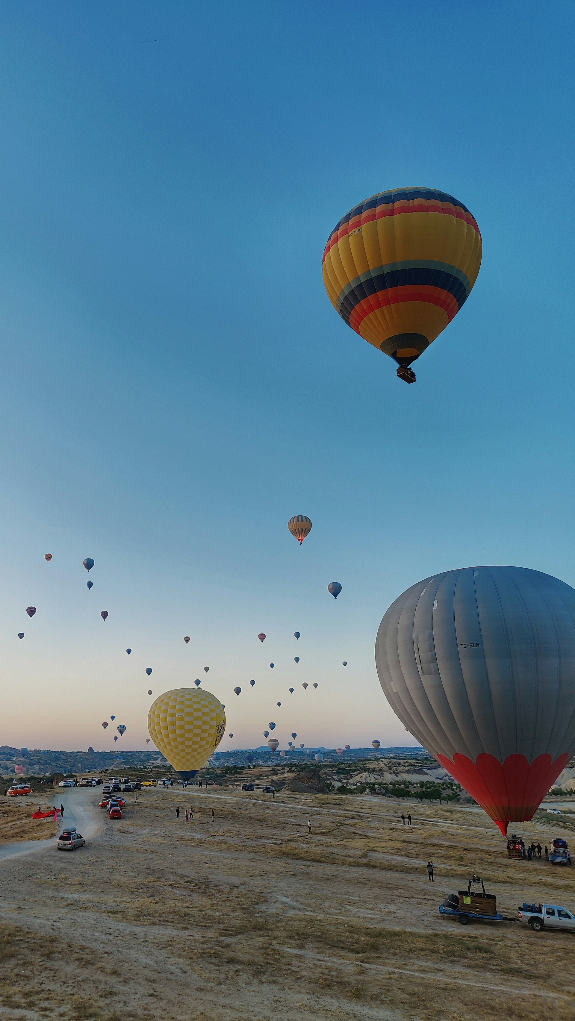 Numerous hot air balloons float majestically over a vast open landscape. The sky is clear with a gentle gradient transitioning from light to deep blue. Several vehicles and individuals are visible on the ground, likely preparing for or observing the balloon flight. The scene conveys a sense of adventure and tranquility.