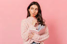 a woman in a pink sweater is holding a book and looking at the camera