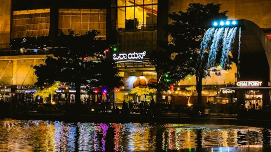 A vibrant cityscape at night featuring a plaza with illuminated signs and buildings. A waterfall installation is prominently displayed next to a tree, with colorful lights and reflections visible in the water below. Crowds of people are gathered around various food establishments, creating a lively atmosphere.