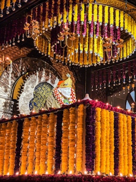 An ornate structure decorated with vibrant marigold garlands in shades of yellow and purple surrounds a statue. Above, chandeliers add to the grandeur with lights reflecting off metallic surfaces. The background features an intricately designed arch with silver-toned details.