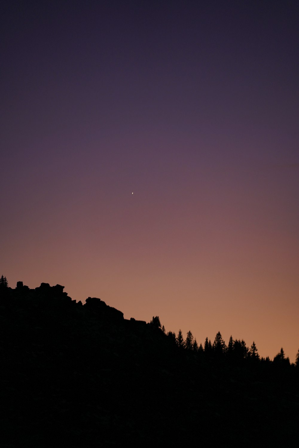 the moon is setting over a hill with trees in the foreground