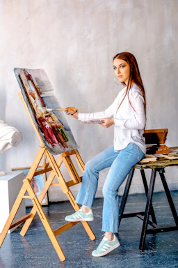 A woman is seated on a stool in a studio, painting on a canvas positioned on an easel. She is wearing a white shirt, blue jeans, and sneakers, and has long brown hair. The studio has a soft, neutral-toned wall, and there is an arrangement of art supplies and materials nearby.