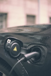 A close-up of an electric vehicle charging port with a charging cable plugged in. The vehicle appears to be black, with raindrops visible on its surface, suggesting it is outdoors in rainy weather. The charging port cover is open, revealing a circular design with a yellow caution symbol.