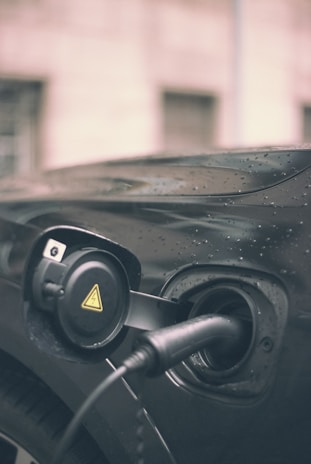 A close-up of an electric vehicle charging port with a charging cable plugged in. The vehicle appears to be black, with raindrops visible on its surface, suggesting it is outdoors in rainy weather. The charging port cover is open, revealing a circular design with a yellow caution symbol.